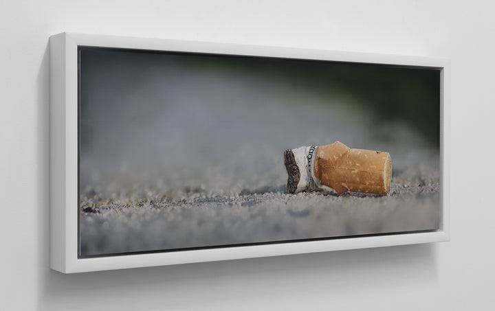 Simon Taylor, Kiss Me Goodbye, oil and acrylic on aluminium, 25 x 60cm – photorealist painting of a discarded cigarette butt lying on gritty ground, with detailed ash, torn paper and shallow depth of field in muted grey and brown tones.