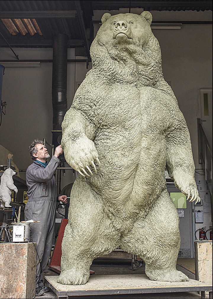 Monumental bronze sculpture of a life-size Kodiak brown bear standing upright on a large plinth inside an industrial foundry setting. The bear’s powerful muscular form, dense fur texture, and extended claws are rendered with extreme realism, conveying scale, strength, and dominance, while a human figure at the base provides dramatic contrast to emphasise the sculpture’s immense height and physical presence.
