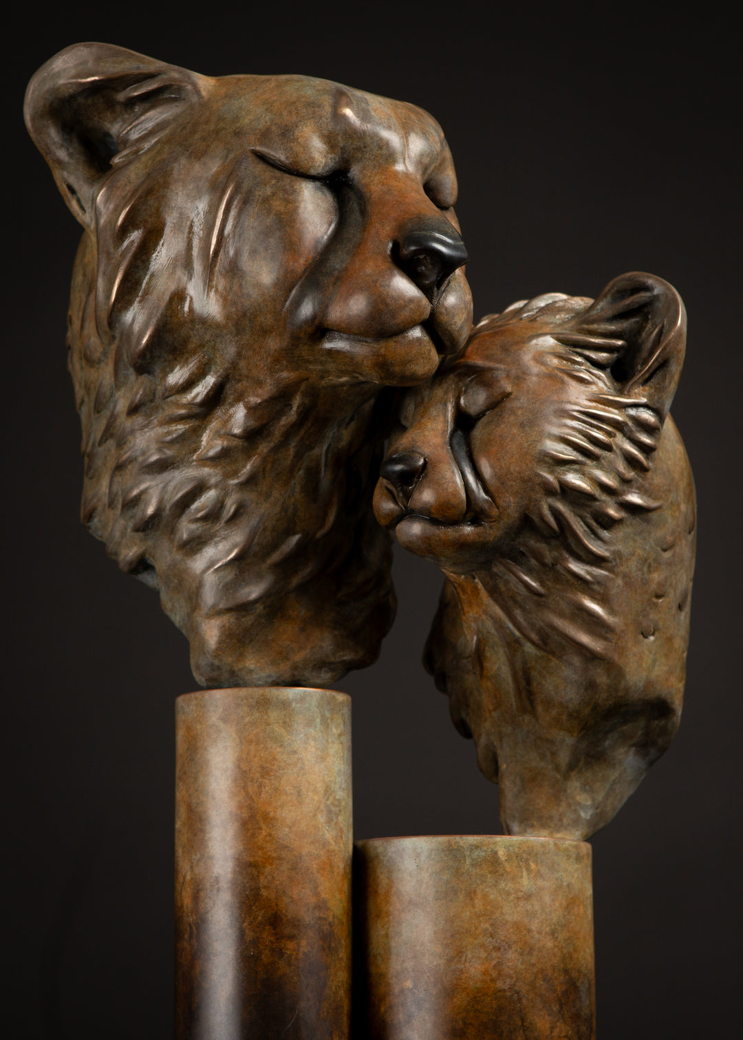 Kulina cheetah mother and cub bronze sculpture by Min Reid, tender wildlife sculpture showing a protective cheetah nuzzling her cub, warm golden brown bronze patina mounted on tall cylindrical bronze plinths against a dark background