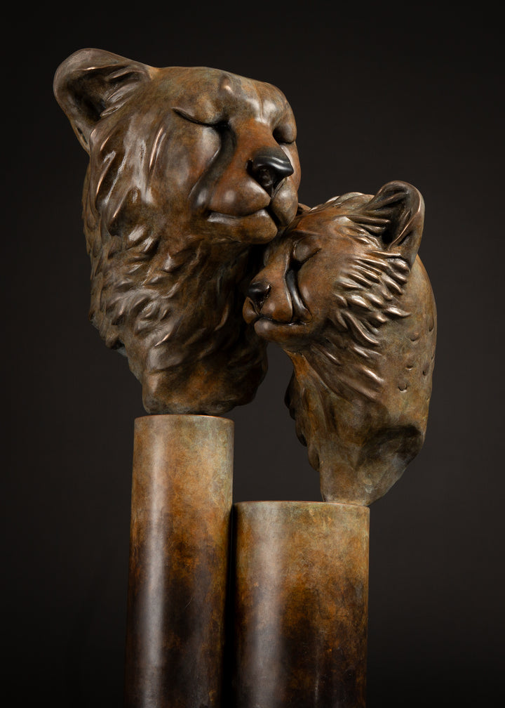 Kulina cheetah mother and cub bronze sculpture by Min Reid, tender wildlife sculpture showing a protective cheetah nuzzling her cub, warm golden brown bronze patina mounted on tall cylindrical bronze plinths against a dark background
