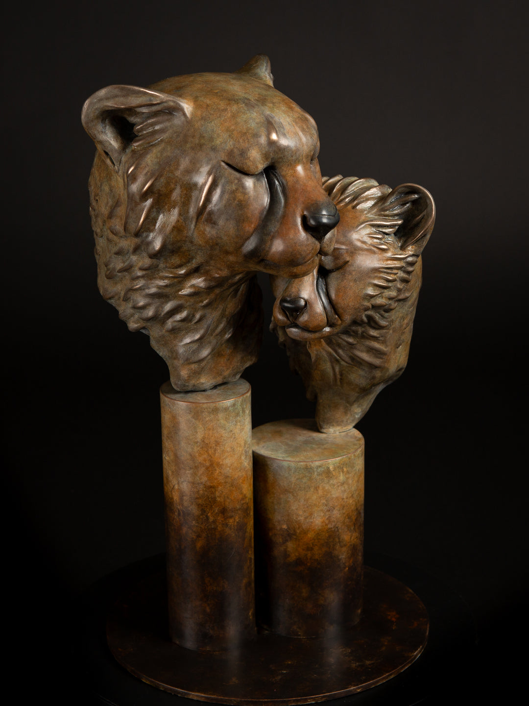 Kulina cheetah mother and cub bronze sculpture by Min Reid, tender wildlife sculpture showing a protective cheetah nuzzling her cub, warm golden brown bronze patina mounted on tall cylindrical bronze plinths against a dark background