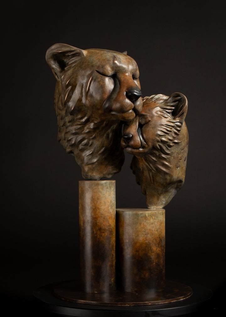Kulina cheetah mother and cub bronze sculpture by Min Reid, tender wildlife sculpture showing a protective cheetah nuzzling her cub, warm golden brown bronze patina mounted on tall cylindrical bronze plinths against a dark background