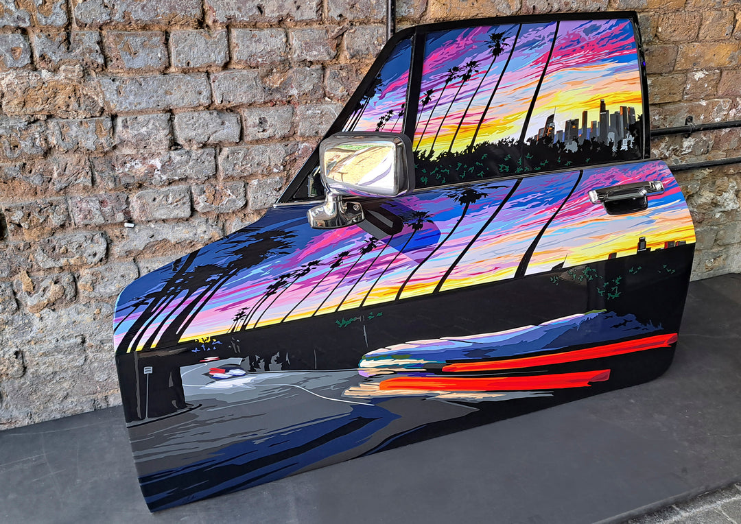Hand-cut vinyl artwork by Joel Clark on a 1980s Jaguar XJ-S door, depicting a vibrant Los Angeles sunset with palm trees, a winding road, and city skyline—rendered in bold, stylised layers of colour with a glossy clear-coat finish.