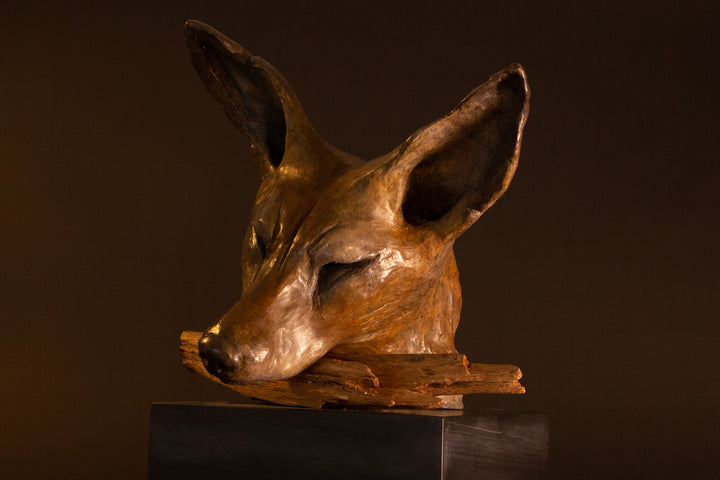 Lean On Me wild dog bronze sculpture by Min Reid, expressive resting wild dog head leaning on a natural wood branch with eyes closed, rich warm bronze patina mounted on a dark plinth against a deep shadowed background