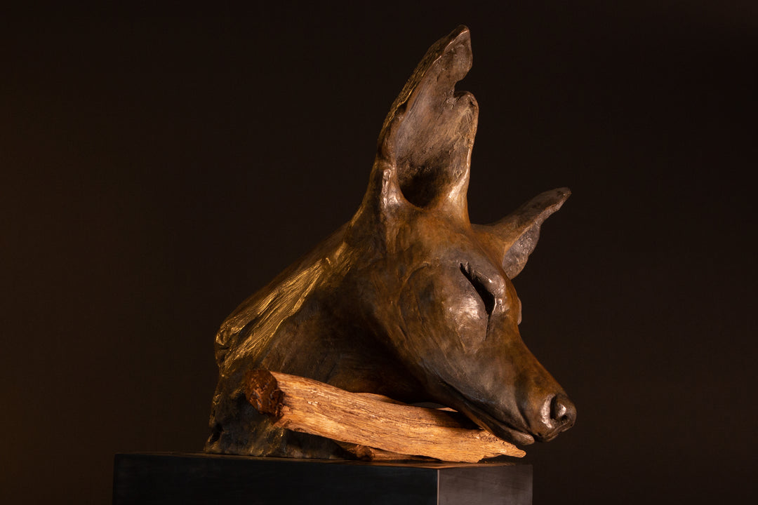 Lean On Me wild dog bronze sculpture by Min Reid, expressive resting wild dog head leaning on a natural wood branch with eyes closed, rich warm bronze patina mounted on a dark plinth against a deep shadowed background