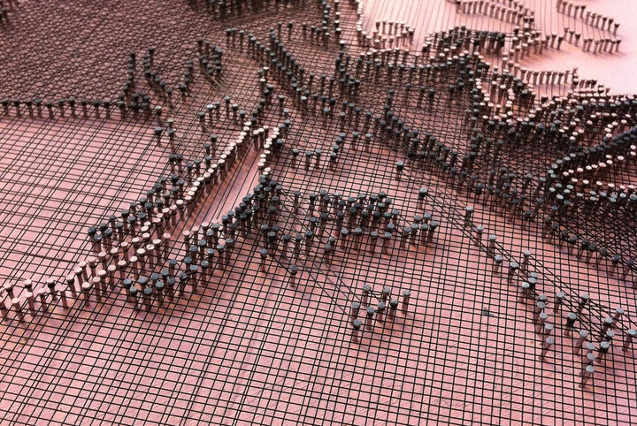 Portrait of Lady Gaga constructed from fine copper wire tensioned around numerous small nails on a painted board, with layered linear wiring forming facial features, hair and hat through tonal variation, depth and shadow across a textured pink-toned surface.