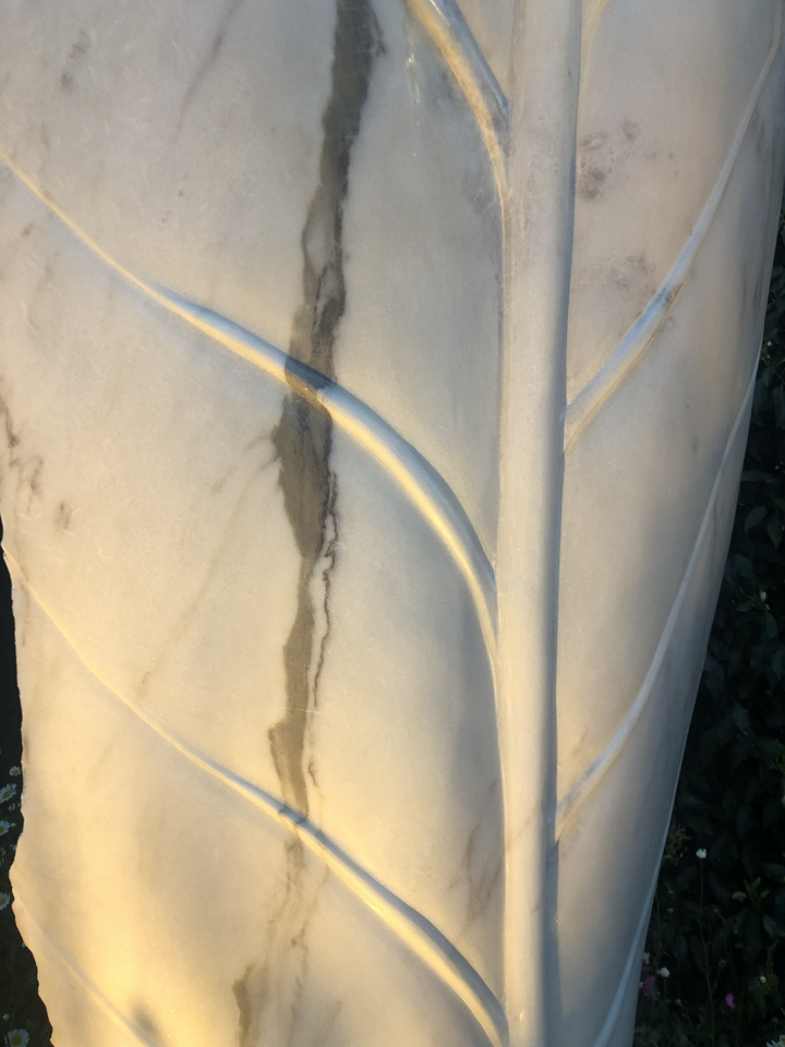 ‘Guardians’ by Nicolas Moreton MRSS, a pair of unique contemporary botanical stone sculptures carved from pale white Portuguese Marble. The first sculpture features a tall, upright leaf with a smooth surface and finely carved central vein and branching leaf veins. The second features an elongated, pointed form with rough natural stone edges, a deeply recessed smooth centre and a raised, textured seed-like structure. Both sculptures stand 260cm tall and are photographed together in a landscaped garden surrou