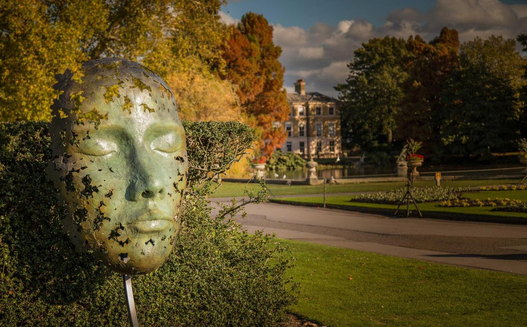 Simon Gudgeon bronze sculpture Leaf Spirit featuring a serene monumental face formed with organic leaf patterns displayed within a landscaped garden and woodland setting.