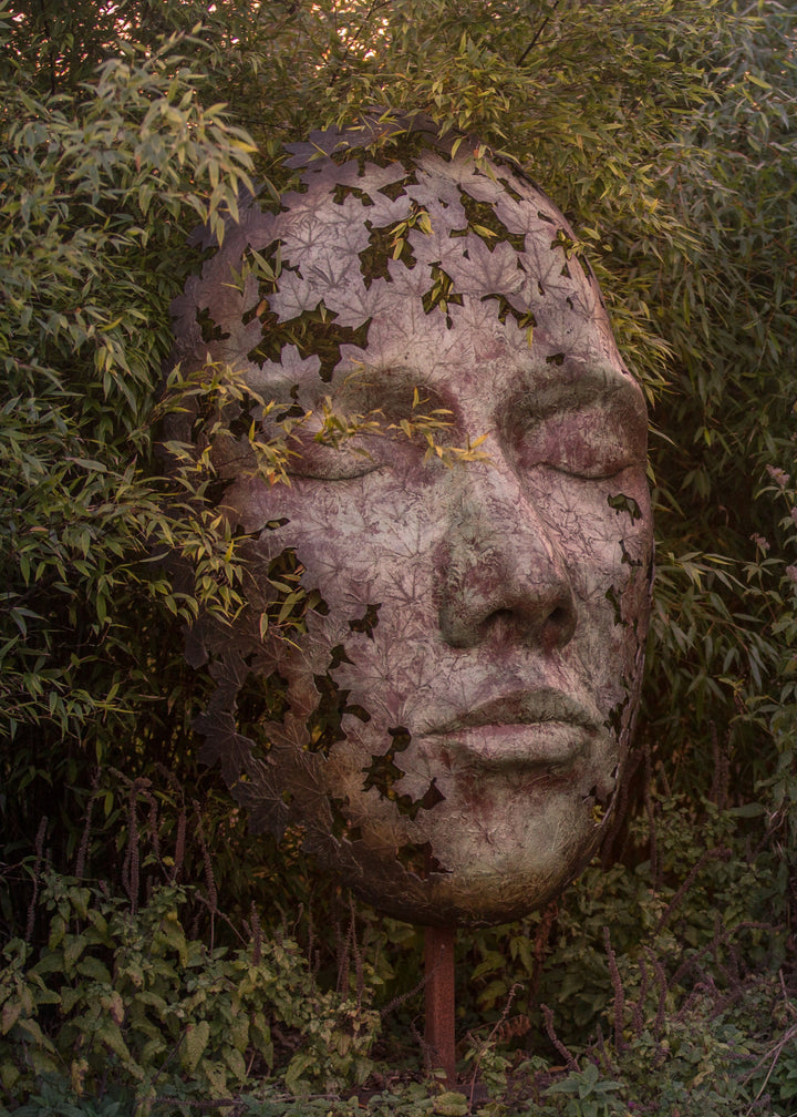 Simon Gudgeon bronze sculpture Leaf Spirit featuring a serene monumental face formed with organic leaf patterns displayed within a landscaped garden and woodland setting.