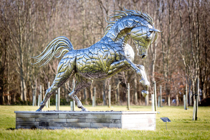 ‘Leaping Horse’ by Michael Turner, a unique, one-of-one stainless steel equestrian sculpture depicting a magnificent life-size horse leaping forward with its front legs raised and hind legs extended. Handcrafted from marine-grade stainless steel, the sculpture features intricately welded metallic plates, a powerful muscular body, flowing mane and sweeping tail. Its polished silver finish reflects the surrounding landscape, while the dynamic pose captures the strength, elegance and movement of a horse in ful