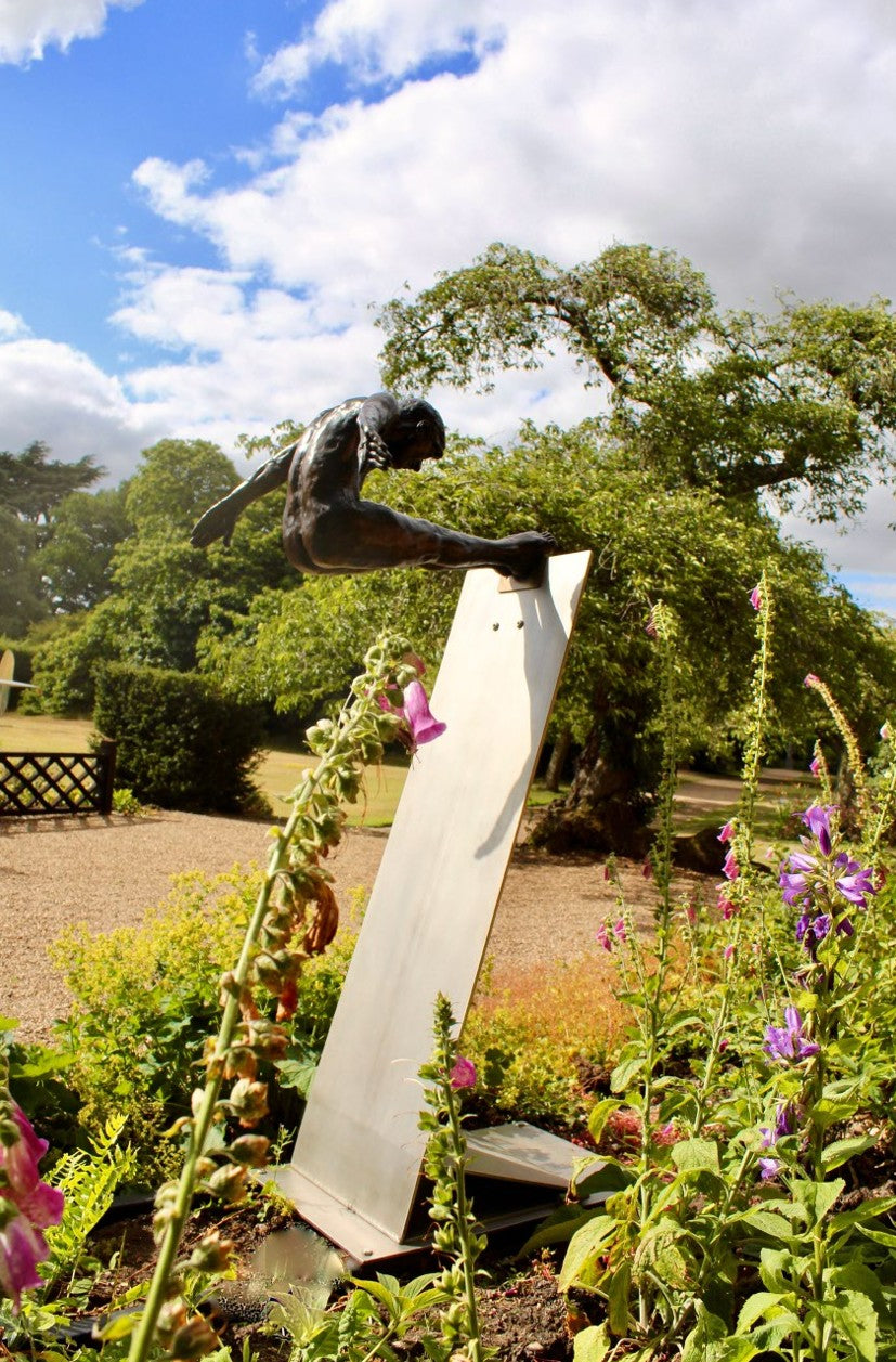 Limited edition bronze sculpture titled Leap of Faith depicting a powerful male figure frozen in mid-flight, arms swept back and legs fully extended forward in a gymnast-like pike position. The muscular form is highly detailed, with realistic anatomy and a burnished bronze finish, mounted on an angled stainless steel platform that suggests a diving board or launching point. Set against open sky or landscaped grounds, the piece captures the moment of total commitment and trust—symbolising courage