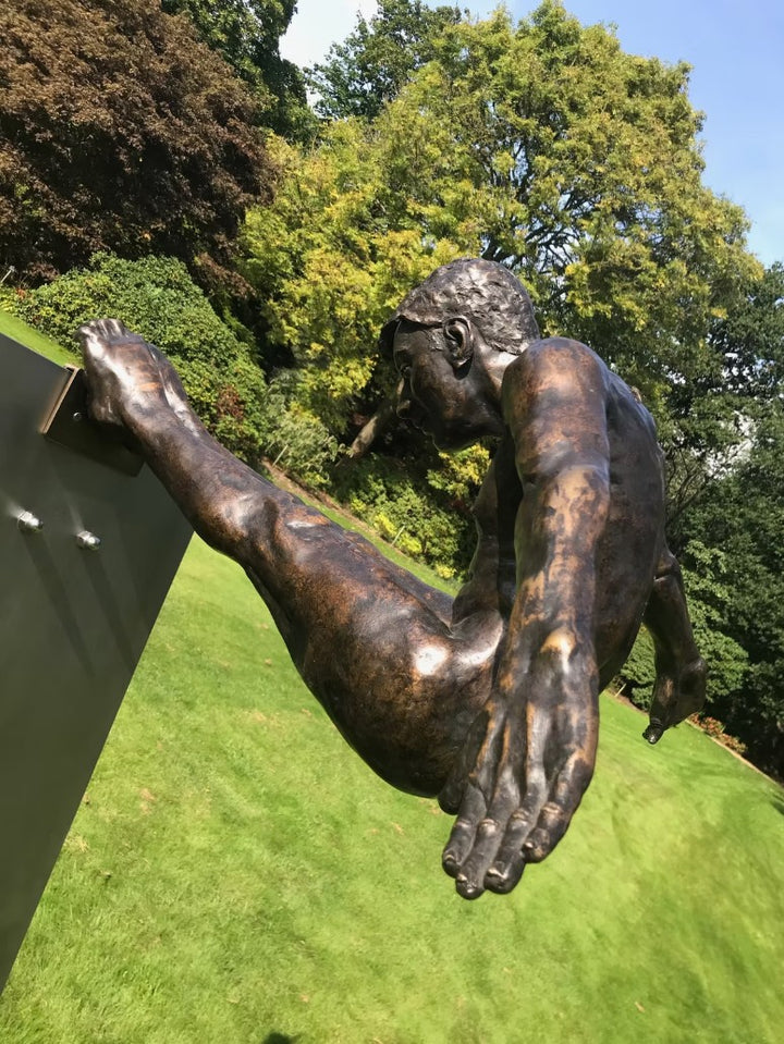 Limited edition bronze sculpture titled Leap of Faith depicting a powerful male figure frozen in mid-flight, arms swept back and legs fully extended forward in a gymnast-like pike position. The muscular form is highly detailed, with realistic anatomy and a burnished bronze finish, mounted on an angled stainless steel platform that suggests a diving board or launching point. Set against open sky or landscaped grounds, the piece captures the moment of total commitment and trust—symbolising courage
