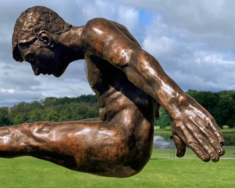 Limited edition bronze sculpture titled Leap of Faith depicting a powerful male figure frozen in mid-flight, arms swept back and legs fully extended forward in a gymnast-like pike position. The muscular form is highly detailed, with realistic anatomy and a burnished bronze finish, mounted on an angled stainless steel platform that suggests a diving board or launching point. Set against open sky or landscaped grounds, the piece captures the moment of total commitment and trust—symbolising courage