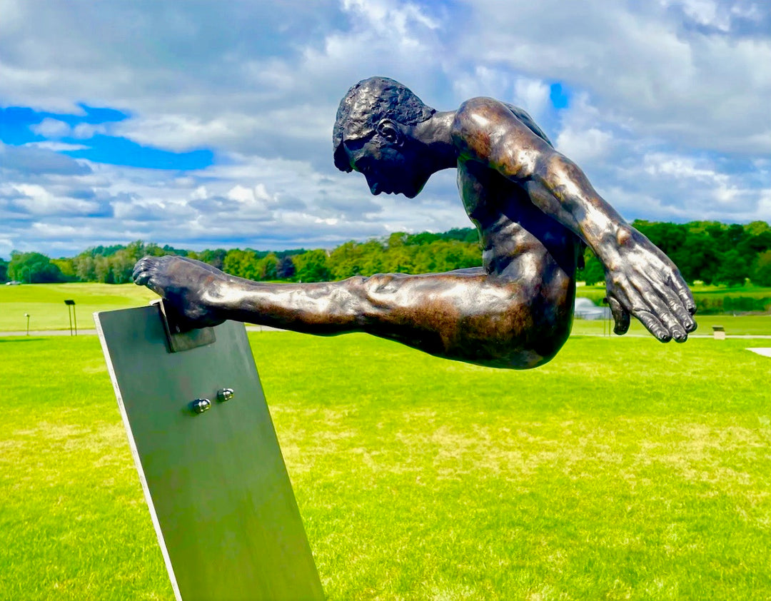 Limited edition bronze sculpture titled Leap of Faith depicting a powerful male figure frozen in mid-flight, arms swept back and legs fully extended forward in a gymnast-like pike position. The muscular form is highly detailed, with realistic anatomy and a burnished bronze finish, mounted on an angled stainless steel platform that suggests a diving board or launching point. Set against open sky or landscaped grounds, the piece captures the moment of total commitment and trust—symbolising courage