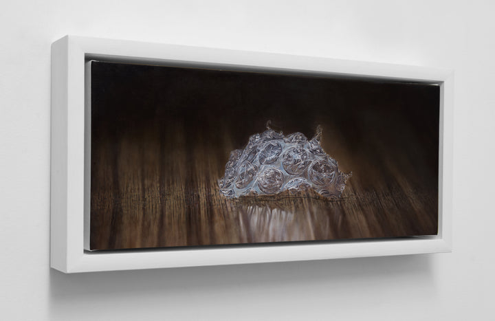 Simon Taylor, Left Behind, 2024, oil on canvas, 17 x 41cm – photorealist painting of a torn fragment of clear bubble wrap resting on a dark wooden surface, with detailed translucent pockets and soft reflections in warm brown light.