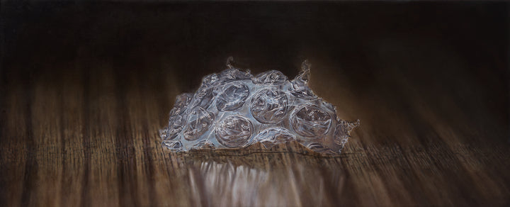 Simon Taylor, Left Behind, 2024, oil on canvas, 17 x 41cm – photorealist painting of a torn fragment of clear bubble wrap resting on a dark wooden surface, with detailed translucent pockets and soft reflections in warm brown light.