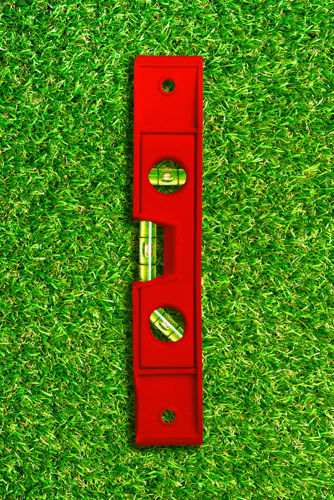 Photograph of a red spirit level placed vertically on a vivid green grass background, combining industrial precision with playful colour contrast.