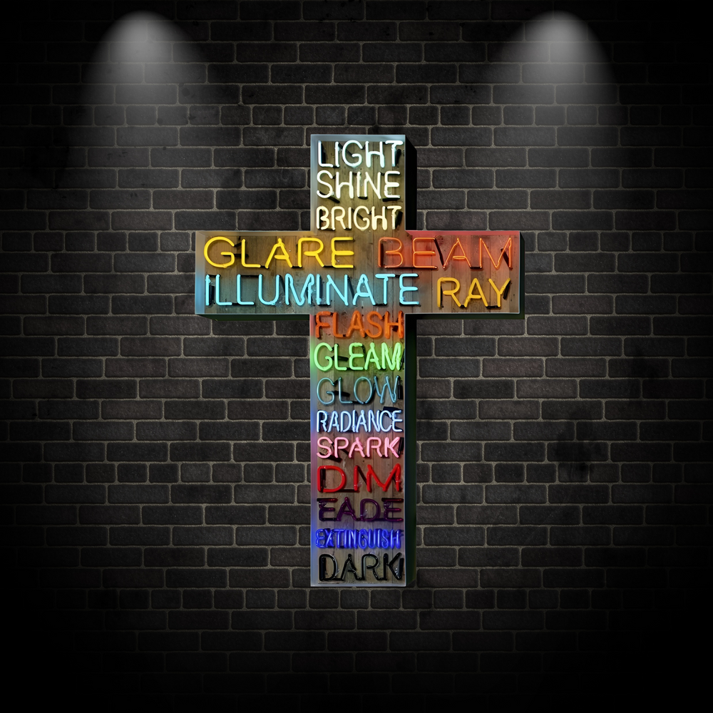 Neon artwork in the shape of a cross by Matthew Bracey, featuring a cascading list of light-related words, from “LIGHT” and “SHINE” at the top to “DARK” and “EXTINGUISH” at the base. Each word is rendered in glowing neon across a spectrum of colours including white, yellow, red, orange, green, blue, pink, and black, mounted on a wooden backdrop.