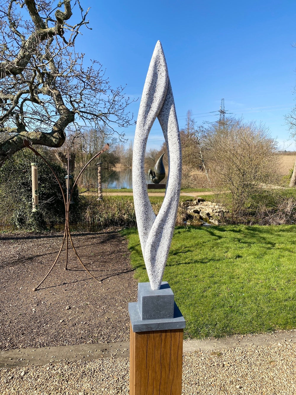 Abstract sculpture Lighthouse – 2024 by Hamish Holman, carved from Cornish granite into a tall, slender vertical form with a central opening and twisting contours, mounted on a dark stone base and oak plinth, displayed outdoors in natural light.