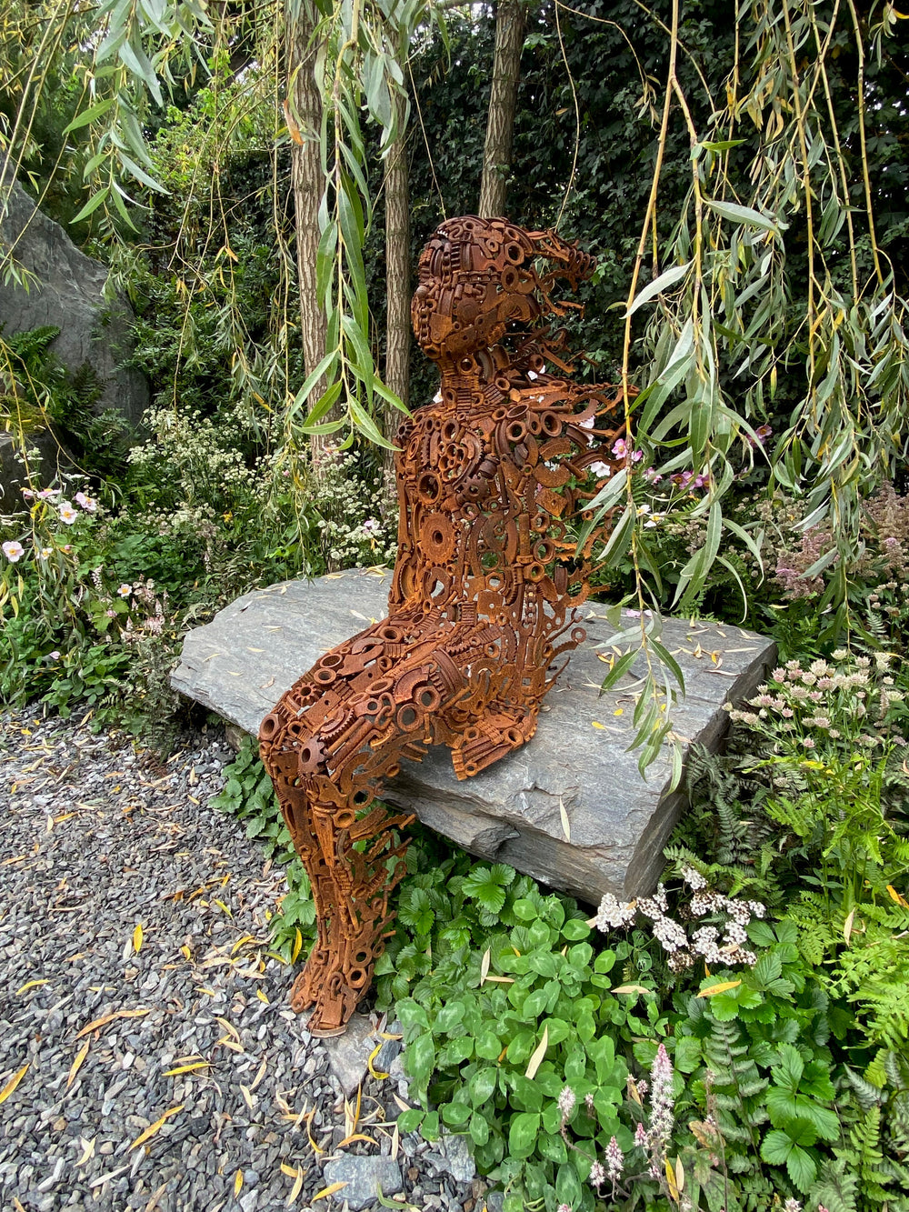 Little Boy by Penny Hardy — a limited edition sculpture formed from intricately welded recycled metal parts, depicting a seated child figure in a moment of stillness, placed on a stone slab within a lush garden setting.