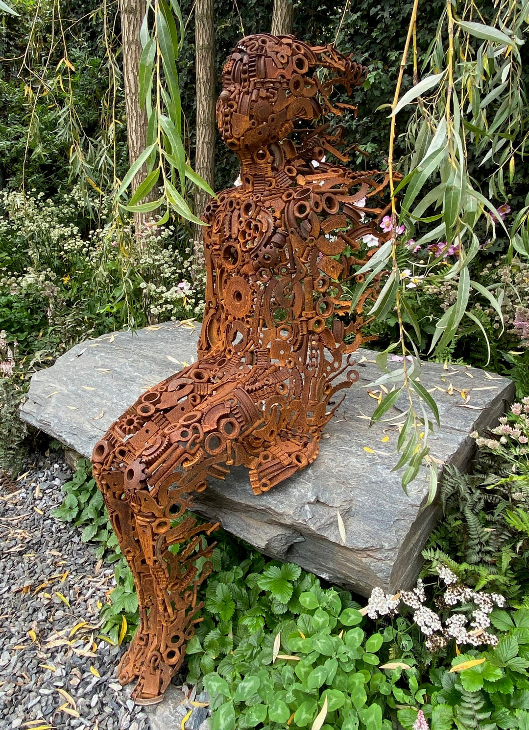 Little Boy by Penny Hardy — a limited edition sculpture formed from intricately welded recycled metal parts, depicting a seated child figure in a moment of stillness, placed on a stone slab within a lush garden setting.