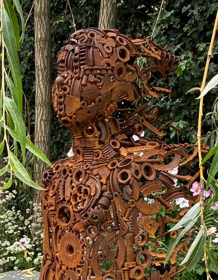 Little Boy by Penny Hardy — a limited edition sculpture formed from intricately welded recycled metal parts, depicting a seated child figure in a moment of stillness, placed on a stone slab within a lush garden setting.