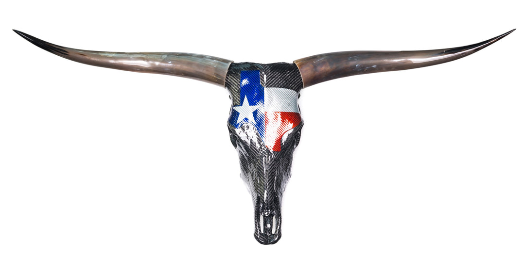 Wall-mounted sculpture of a real Texas Longhorn steer skull wrapped in glossy black carbon fibre with a bold Texan flag design in red, white, and blue Texalium across the forehead. The original curved horns are polished to a brown-black gradient, while titanium nose fittings and colour-coated teeth add Formula 1 detailing. A striking fusion of nature, motorsport, and modern material engineering.
