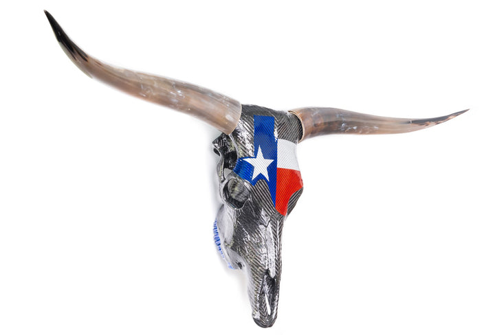 Wall-mounted sculpture of a real Texas Longhorn steer skull wrapped in glossy black carbon fibre with a bold Texan flag design in red, white, and blue Texalium across the forehead. The original curved horns are polished to a brown-black gradient, while titanium nose fittings and colour-coated teeth add Formula 1 detailing. A striking fusion of nature, motorsport, and modern material engineering.
