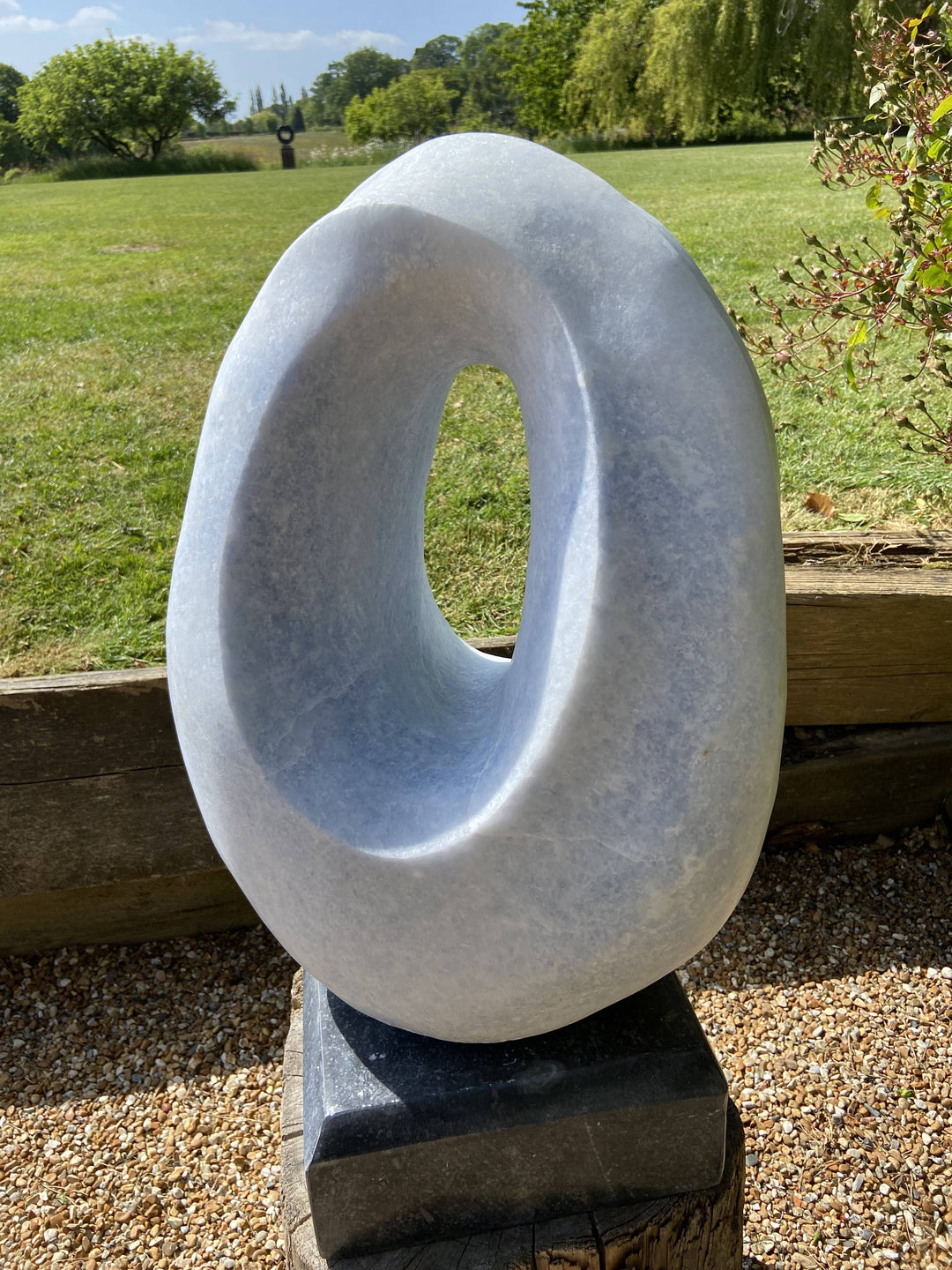 Loop by Brendan Cullen, a smooth hand-carved sculpture in Azure Cielo marble, its elliptical form shaped into a hollowed loop with softly curving edges. The pale blue-grey stone, subtly translucent, captures shifting tones of light, creating a serene, organic presence on a dark stone base.