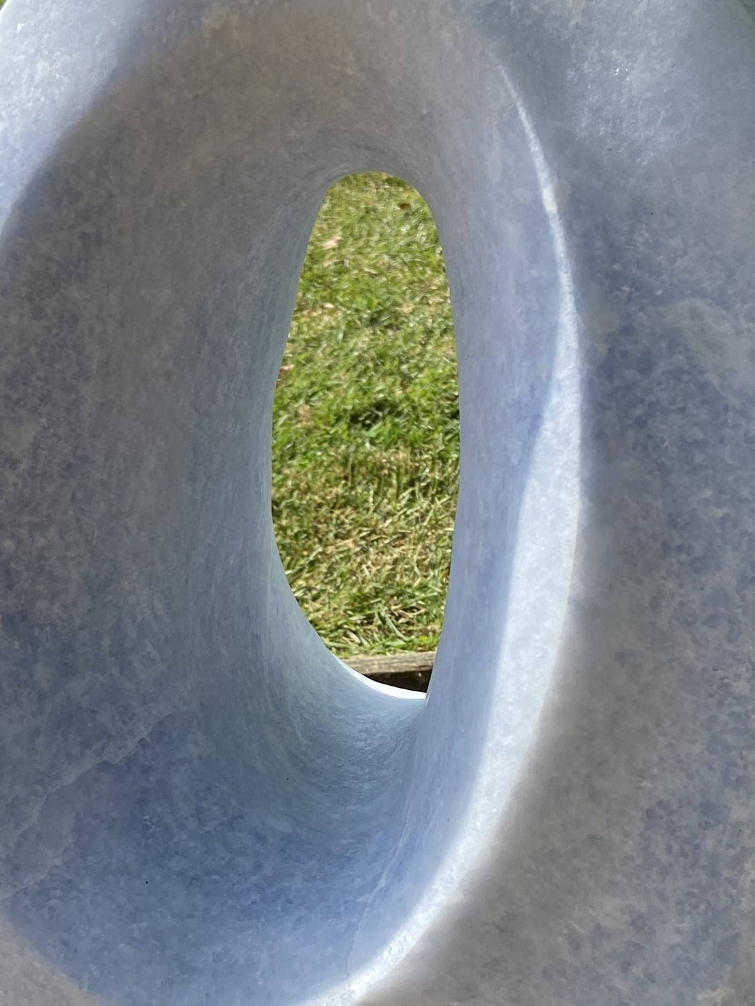 Loop by Brendan Cullen, a smooth hand-carved sculpture in Azure Cielo marble, its elliptical form shaped into a hollowed loop with softly curving edges. The pale blue-grey stone, subtly translucent, captures shifting tones of light, creating a serene, organic presence on a dark stone base.