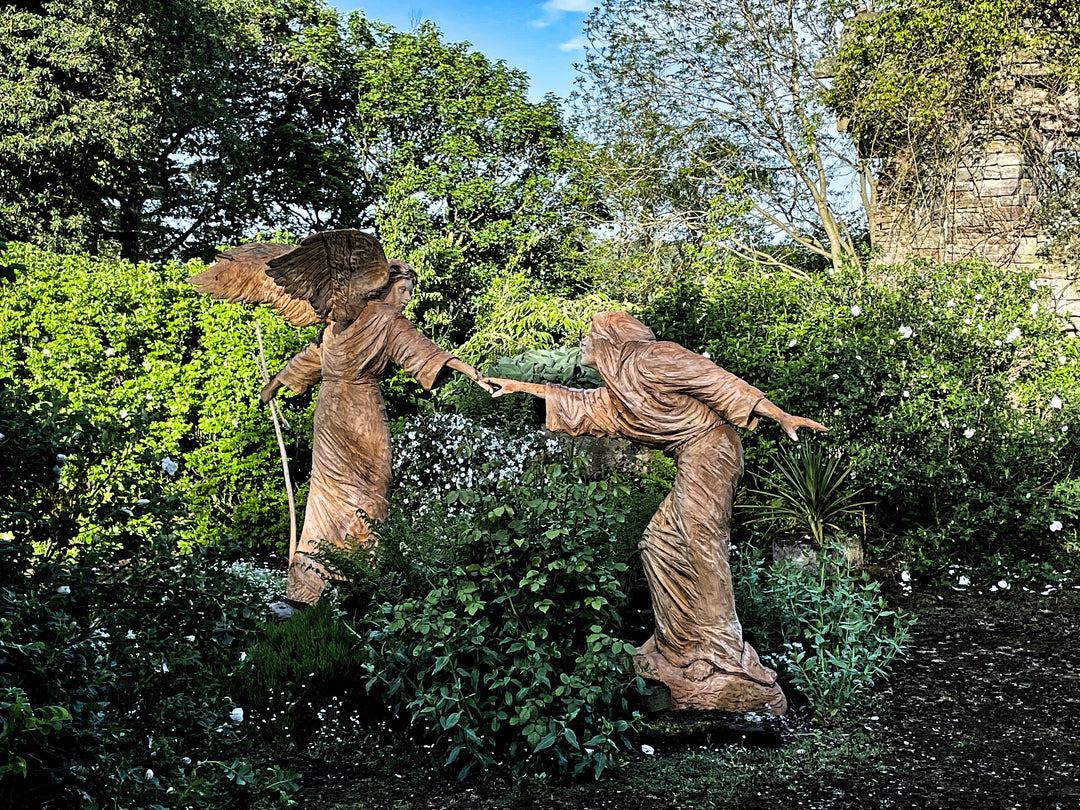 Two life-sized wooden sculptures in a lush garden setting depict a dramatic scene—an angel with outstretched wings reaches toward a hooded, robed figure. Their fingers nearly touch, evoking a moment of divine or spiritual connection. Surrounded by vivid greenery and white blossoms, the earthy tones of the carved figures contrast beautifully with the vibrant natural backdrop.