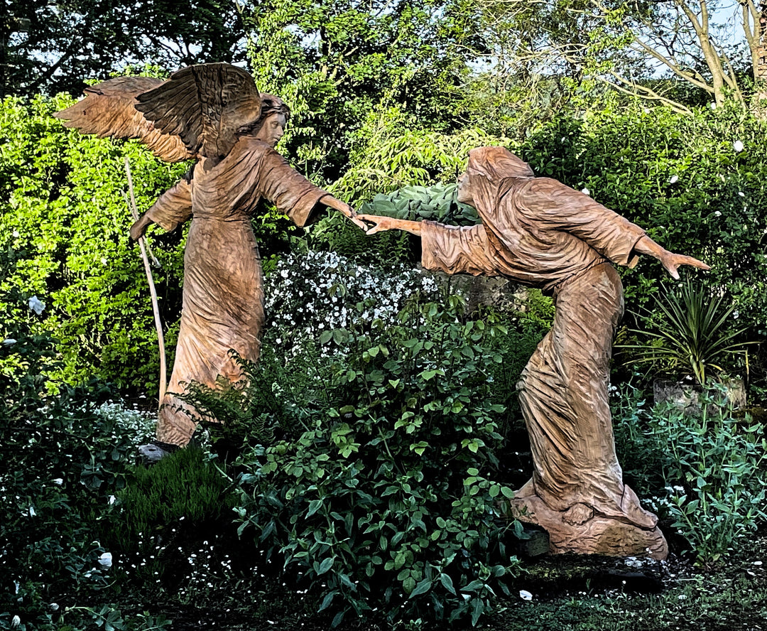 Two life-sized wooden sculptures in a lush garden setting depict a dramatic scene—an angel with outstretched wings reaches toward a hooded, robed figure. Their fingers nearly touch, evoking a moment of divine or spiritual connection. Surrounded by vivid greenery and white blossoms, the earthy tones of the carved figures contrast beautifully with the vibrant natural backdrop.
