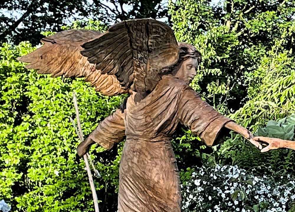 Two life-sized wooden sculptures in a lush garden setting depict a dramatic scene—an angel with outstretched wings reaches toward a hooded, robed figure. Their fingers nearly touch, evoking a moment of divine or spiritual connection. Surrounded by vivid greenery and white blossoms, the earthy tones of the carved figures contrast beautifully with the vibrant natural backdrop.
