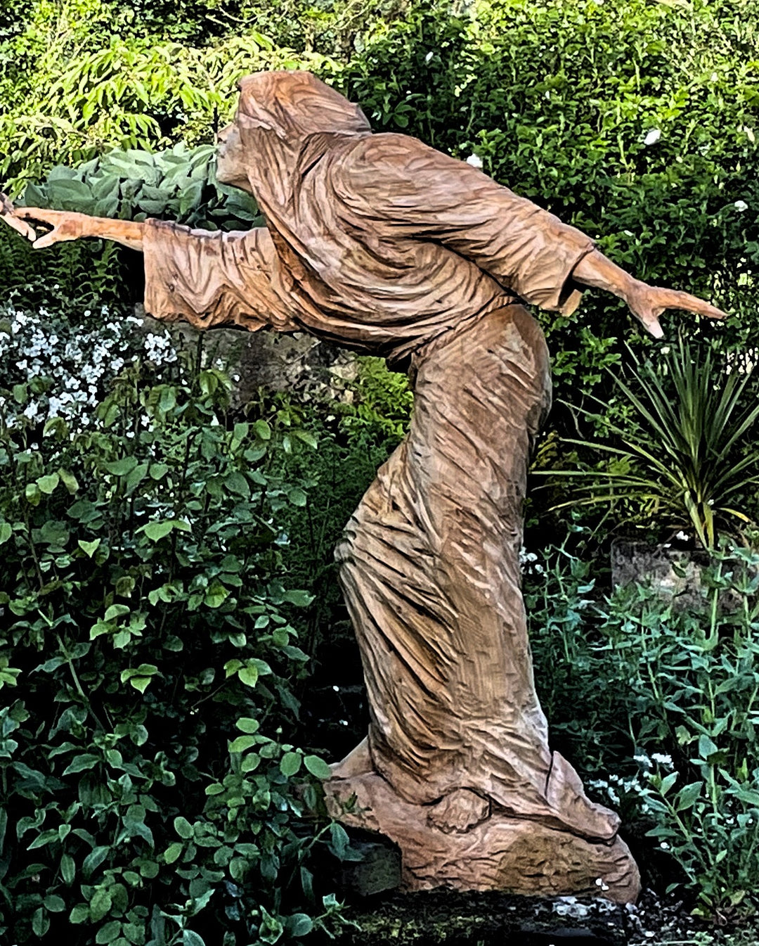 Two life-sized wooden sculptures in a lush garden setting depict a dramatic scene—an angel with outstretched wings reaches toward a hooded, robed figure. Their fingers nearly touch, evoking a moment of divine or spiritual connection. Surrounded by vivid greenery and white blossoms, the earthy tones of the carved figures contrast beautifully with the vibrant natural backdrop.