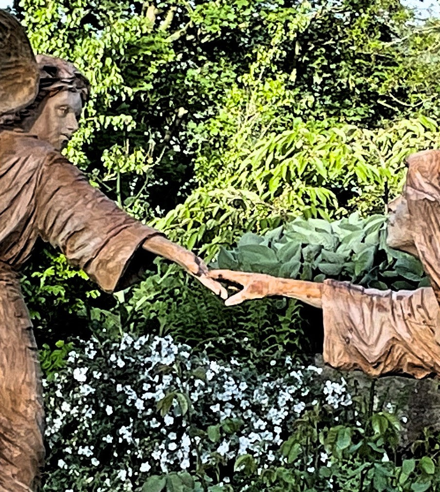 Two life-sized wooden sculptures in a lush garden setting depict a dramatic scene—an angel with outstretched wings reaches toward a hooded, robed figure. Their fingers nearly touch, evoking a moment of divine or spiritual connection. Surrounded by vivid greenery and white blossoms, the earthy tones of the carved figures contrast beautifully with the vibrant natural backdrop.