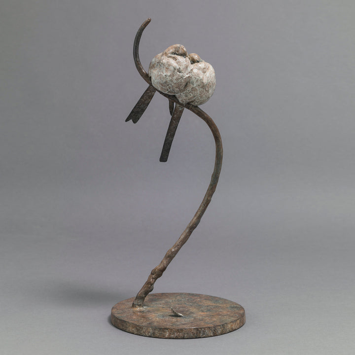 Bronze sculpture of two life-size long-tailed tits nestled closely together on a gently curving branch, their rounded bodies touching as they perch in balance. The birds’ finely textured forms show soft feather detail and subtle patina variations, conveying intimacy, warmth, and quiet stillness against a neutral studio background.