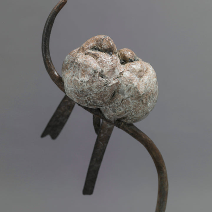 Bronze sculpture of two life-size long-tailed tits nestled closely together on a gently curving branch, their rounded bodies touching as they perch in balance. The birds’ finely textured forms show soft feather detail and subtle patina variations, conveying intimacy, warmth, and quiet stillness against a neutral studio background.
