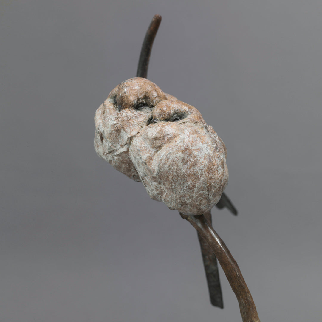 Bronze sculpture of two life-size long-tailed tits nestled closely together on a gently curving branch, their rounded bodies touching as they perch in balance. The birds’ finely textured forms show soft feather detail and subtle patina variations, conveying intimacy, warmth, and quiet stillness against a neutral studio background.