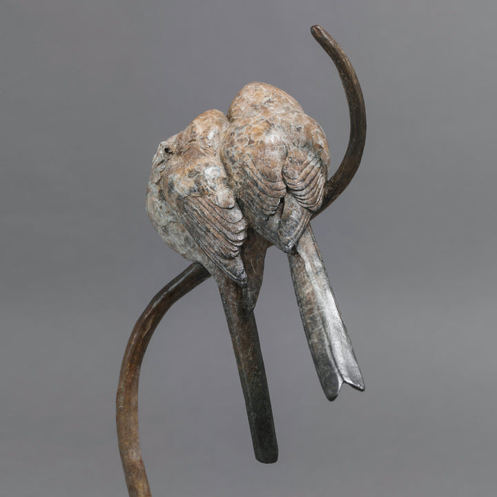 Bronze sculpture of two life-size long-tailed tits nestled closely together on a gently curving branch, their rounded bodies touching as they perch in balance. The birds’ finely textured forms show soft feather detail and subtle patina variations, conveying intimacy, warmth, and quiet stillness against a neutral studio background.