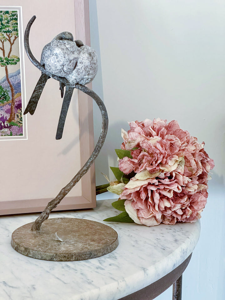 Bronze sculpture of two life-size long-tailed tits nestled closely together on a gently curving branch, their rounded bodies touching as they perch in balance. The birds’ finely textured forms show soft feather detail and subtle patina variations, conveying intimacy, warmth, and quiet stillness against a neutral studio background.