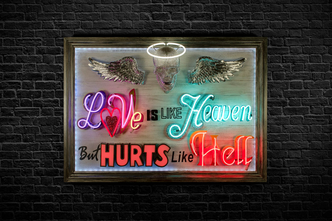 Mixed-media neon artwork by Matthew Bracey featuring the phrase "Love is like Heaven but hurts like Hell." The composition combines vivid neon typography in pink, red, white, blue, and orange, with a central crystal-studded skull and halo, and silver glitter angel wings mounted on a light wood panel.