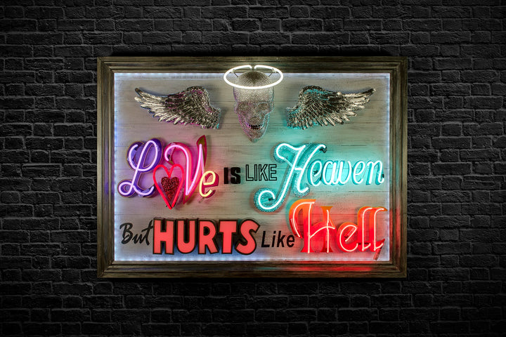 Mixed-media neon artwork by Matthew Bracey featuring the phrase "Love is like Heaven but hurts like Hell." The composition combines vivid neon typography in pink, red, white, blue, and orange, with a central crystal-studded skull and halo, and silver glitter angel wings mounted on a light wood panel.