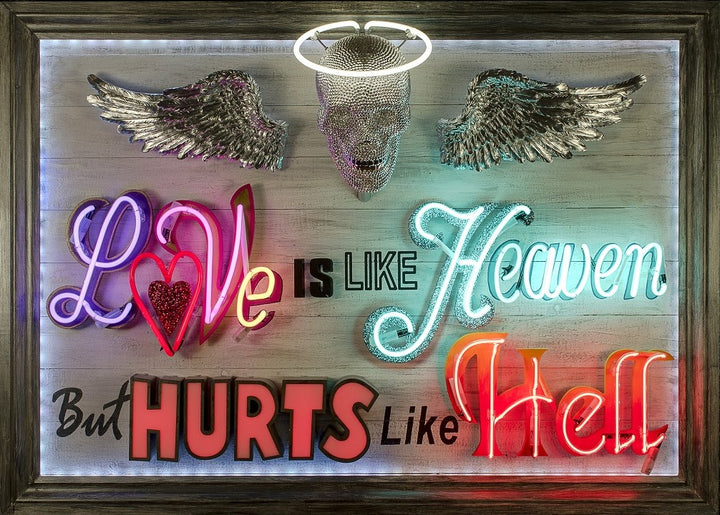 Mixed-media neon artwork by Matthew Bracey featuring the phrase "Love is like Heaven but hurts like Hell." The composition combines vivid neon typography in pink, red, white, blue, and orange, with a central crystal-studded skull and halo, and silver glitter angel wings mounted on a light wood panel.