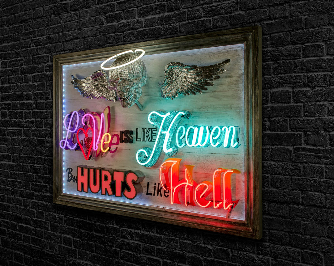 Mixed-media neon artwork by Matthew Bracey featuring the phrase "Love is like Heaven but hurts like Hell." The composition combines vivid neon typography in pink, red, white, blue, and orange, with a central crystal-studded skull and halo, and silver glitter angel wings mounted on a light wood panel.