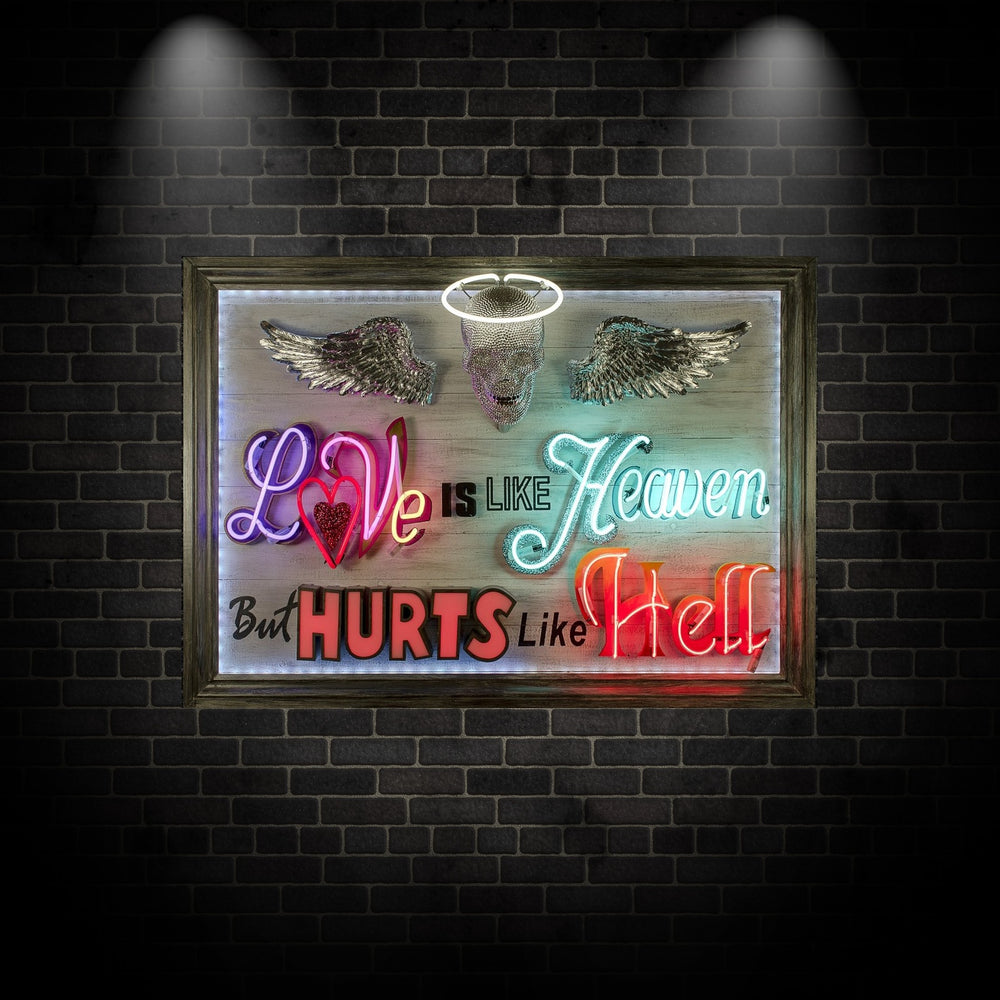 Mixed-media neon artwork by Matthew Bracey featuring the phrase "Love is like Heaven but hurts like Hell." The composition combines vivid neon typography in pink, red, white, blue, and orange, with a central crystal-studded skull and halo, and silver glitter angel wings mounted on a light wood panel.