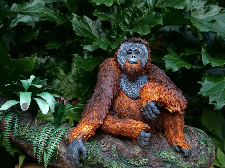 Highly detailed sculpture of a Bornean orangutan lounging on a branch, featuring hand-cut burnt orange paper fur, textured grey-blue skin, and expressive amber eyes, surrounded by lush green tropical foliage.