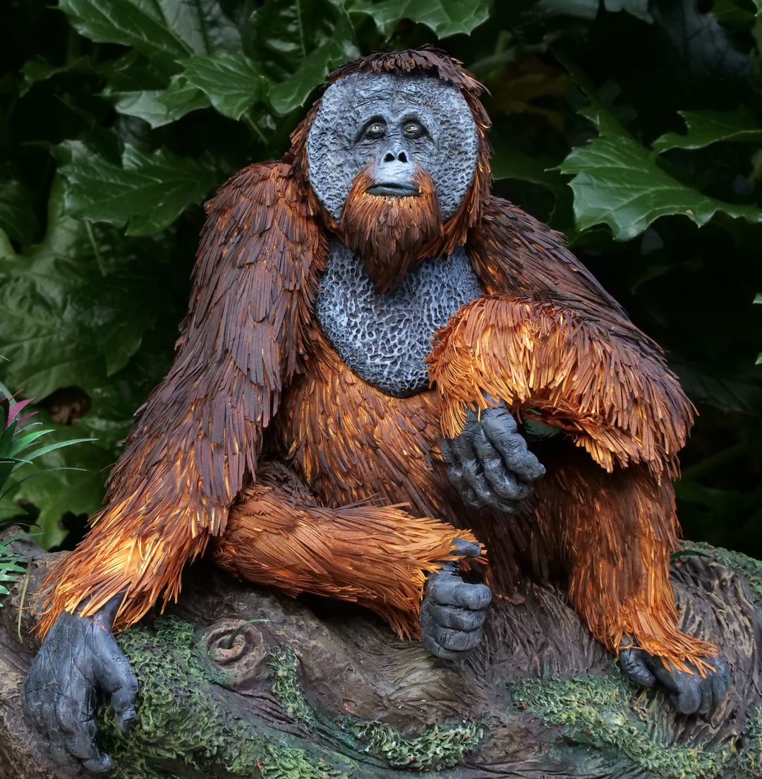 Highly detailed sculpture of a Bornean orangutan lounging on a branch, featuring hand-cut burnt orange paper fur, textured grey-blue skin, and expressive amber eyes, surrounded by lush green tropical foliage.