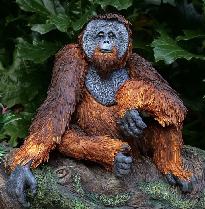 Highly detailed sculpture of a Bornean orangutan lounging on a branch, featuring hand-cut burnt orange paper fur, textured grey-blue skin, and expressive amber eyes, surrounded by lush green tropical foliage.