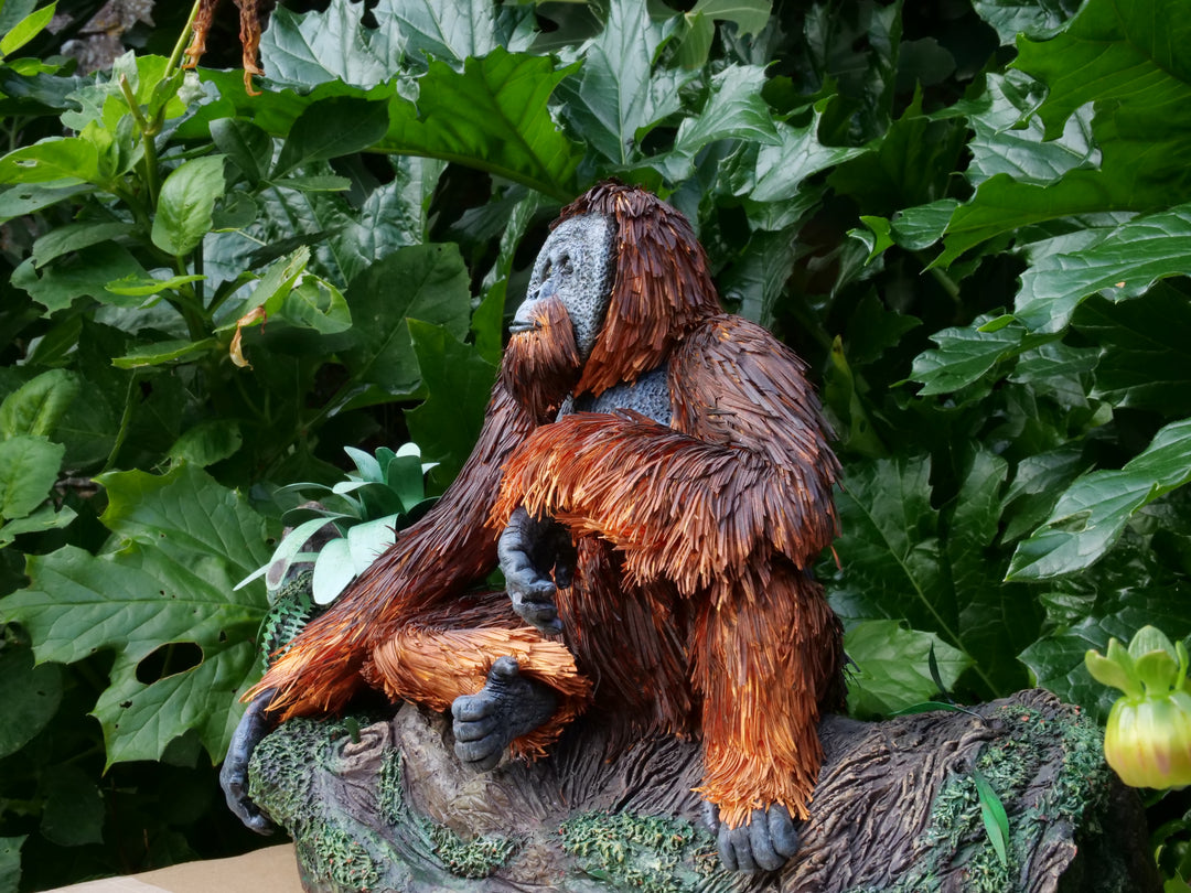 Highly detailed sculpture of a Bornean orangutan lounging on a branch, featuring hand-cut burnt orange paper fur, textured grey-blue skin, and expressive amber eyes, surrounded by lush green tropical foliage.