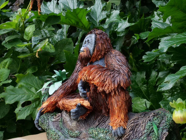 Highly detailed sculpture of a Bornean orangutan lounging on a branch, featuring hand-cut burnt orange paper fur, textured grey-blue skin, and expressive amber eyes, surrounded by lush green tropical foliage.